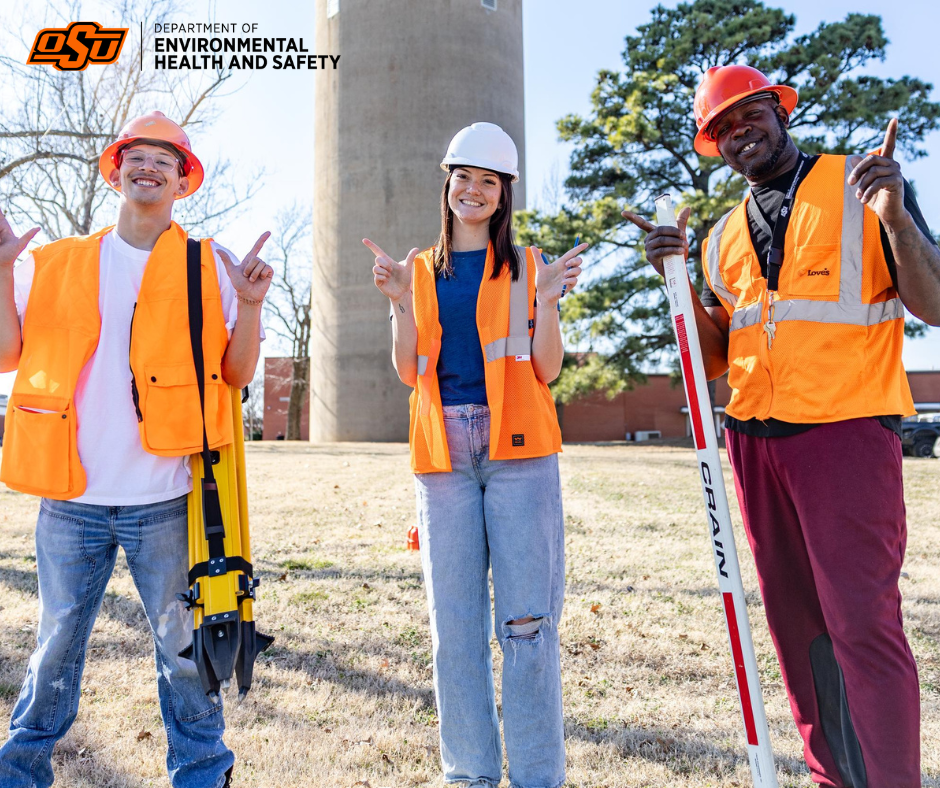 Three people in construction gear stand together and do the "Go Pokes" hand sign. 